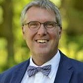 Mann mitte 50er mit Brille und weißem Hemd mit einer bunten Fliege und blauem Sacko vor grünem Naturhindergrund