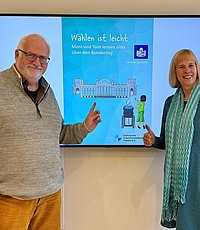 Mann udn Frau stehen vor einem Bildschirm an der Wand wo die Broschüre "Wählen ist leicht" abgebildet ist