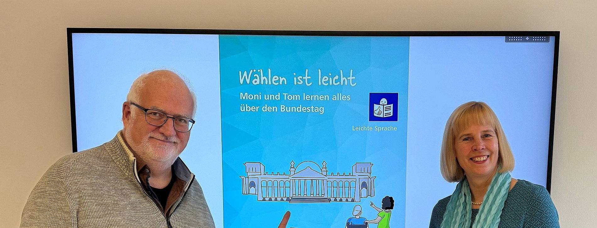 Mann udn Frau stehen vor einem Bildschirm an der Wand wo die Broschüre "Wählen ist leicht" abgebildet ist