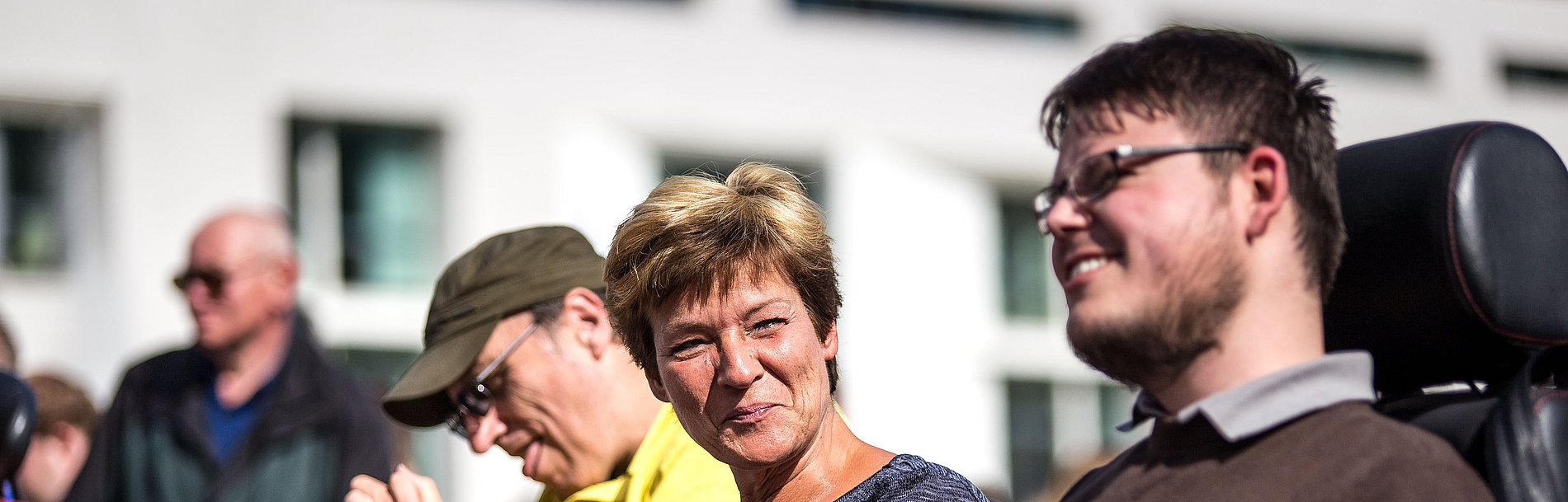 Im vordergrund ist ein Mann mit Brille im Rollstuhl zu sehen, neben ihm sitzt eine Frau und im Hintergrund noch ein Mann mit Kappe