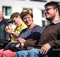 Im vordergrund ist ein Mann mit Brille im Rollstuhl zu sehen, neben ihm sitzt eine Frau und im Hintergrund noch ein Mann mit Kappe