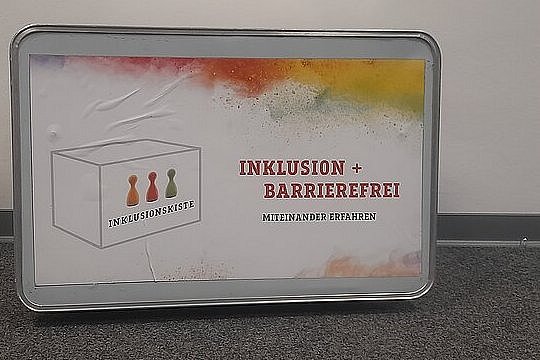 eine Metallkiste mit der Aufschrift Inklusion+Barrierefreiheit drum herum eine Farbwolke und ein Logo 