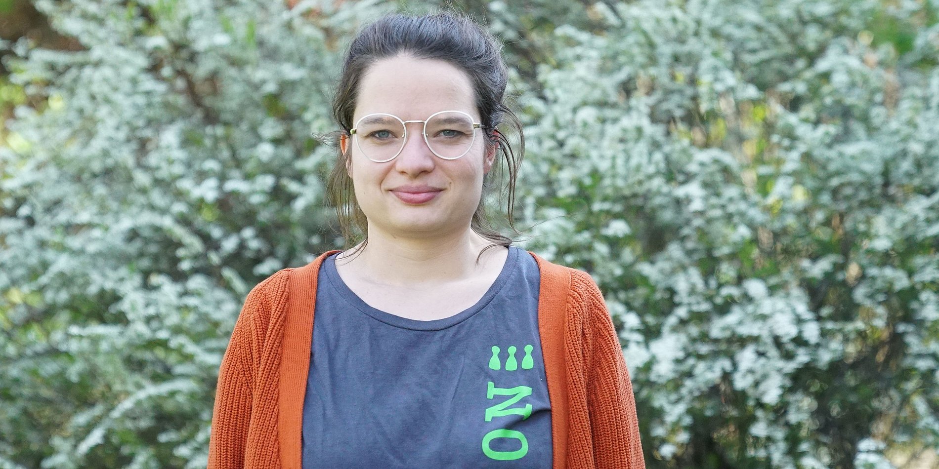 junge Frau mit Brille in einem grauen T-Shirt mit grüner Aufschrift "Inklusion" darüber einer roten Strickjacke vor einem grünen Buschwerk