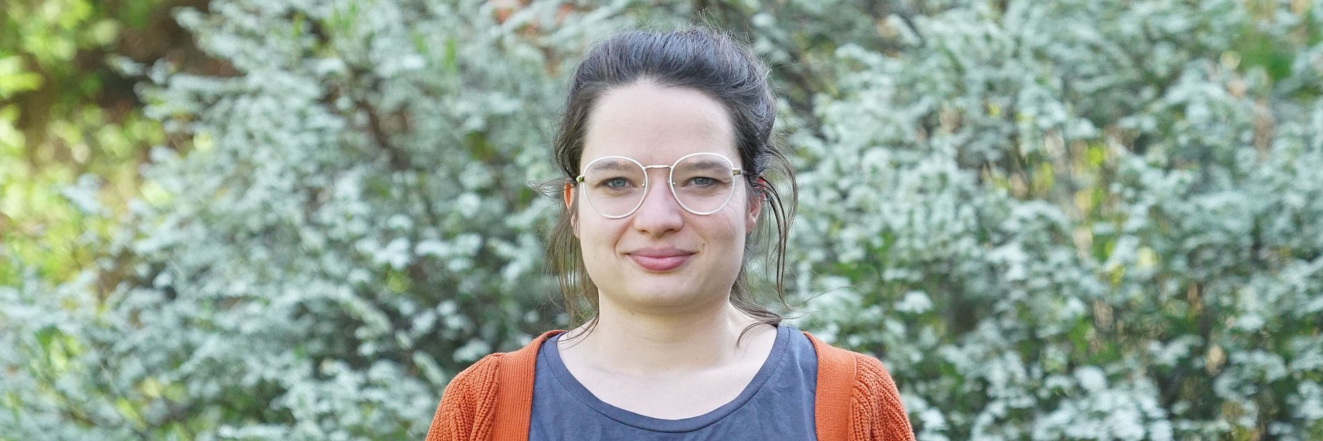 junge Frau mit Brille in einem grauen T-Shirt mit grüner Aufschrift "Inklusion" darüber einer roten Strickjacke vor einem grünen Buschwerk