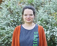 junge Frau an die 30 Jahre mit weiser schmaler Brille grauem T-Shirt, roter Jacke vor grünem Busch