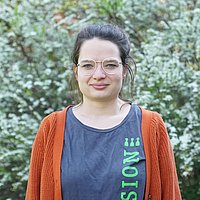 junge Frau mit Brille in einem grauen T-Shirt mit grüner Aufschrift "Inklusion" darüber einer roten Strickjacke vor einem grünen Buschwerk