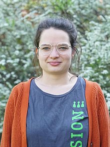 junge Frau mit Brille in einem grauen T-Shirt mit grüner Aufschrift "Inklusion" darüber einer roten Strickjacke vor einem grünen Buschwerk