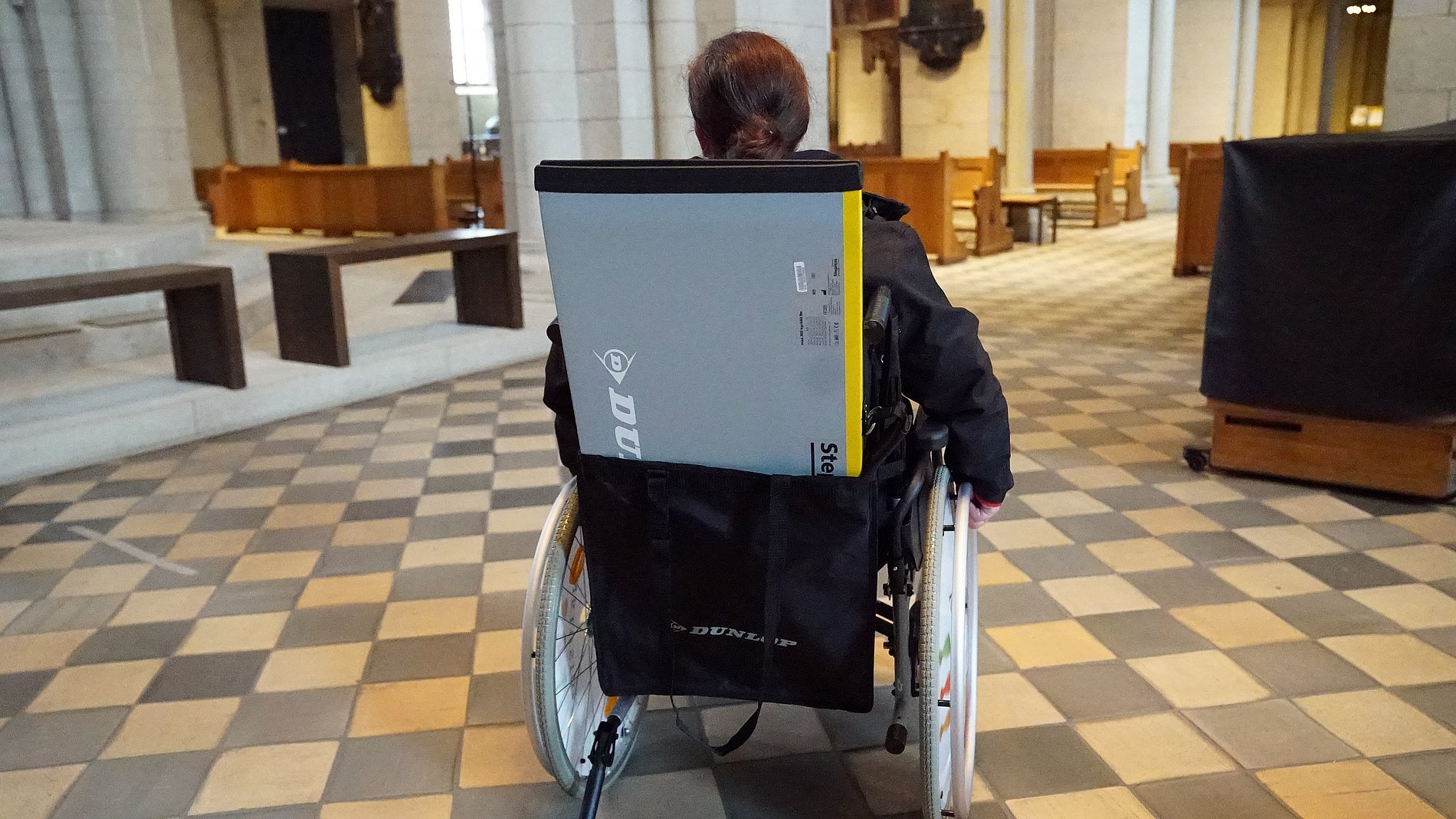 Rollstuhlfahrer von hinten mit einer kleinen Rampe in einer Taswche die am Rollstuhl hängt