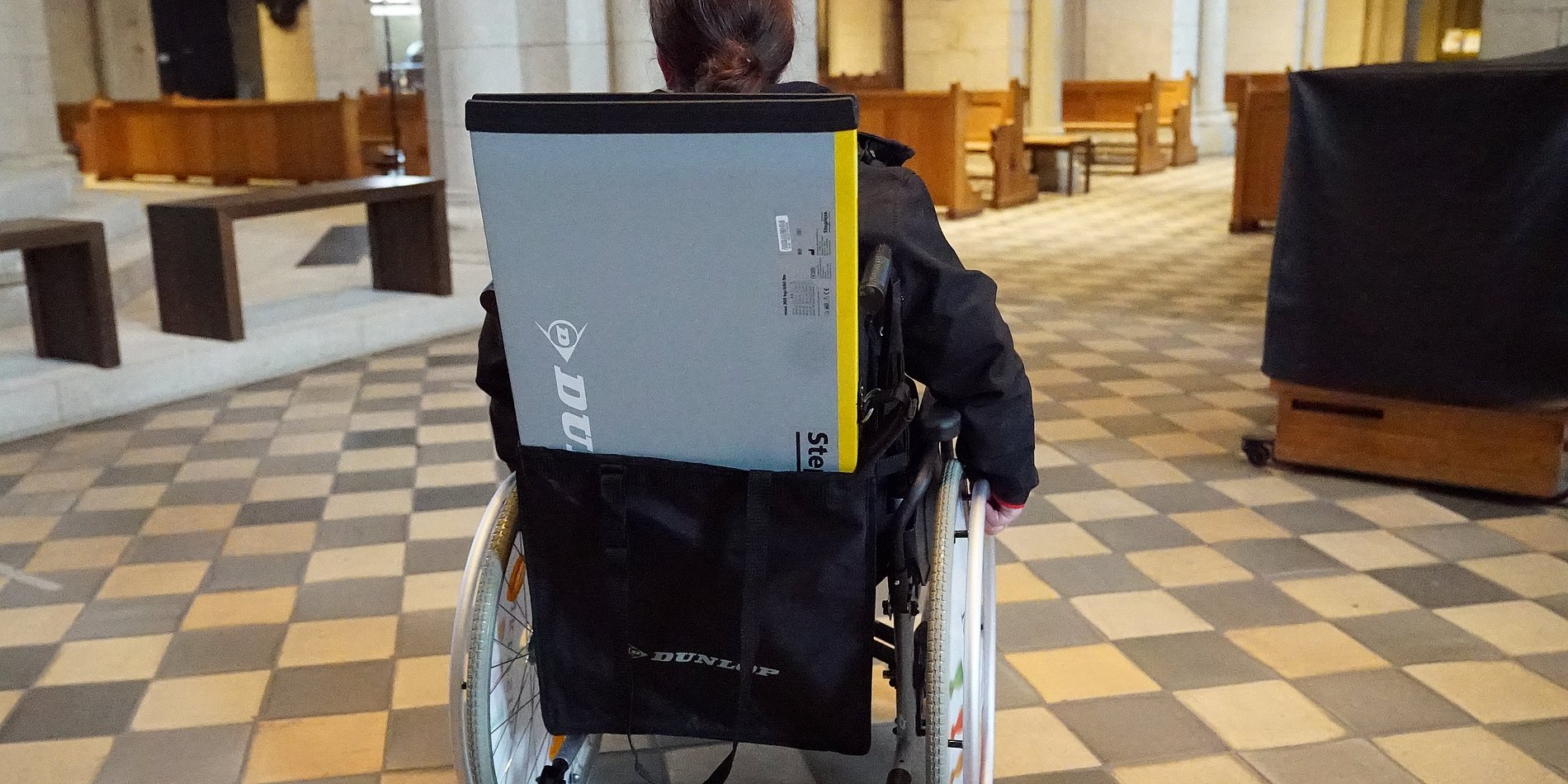 Rollstuhlfahrer von hinten mit einer kleinen Rampe in einer Taswche die am Rollstuhl hängt