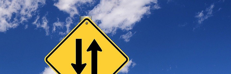 gelbes viereckiges Schild mit zwei entgegengesetzen Pfeilen vor einem blauen Himmel mit Wolken, darunter ein grünes Schild mit der Aufschrift in gelben Buchstaben "Give and Take"