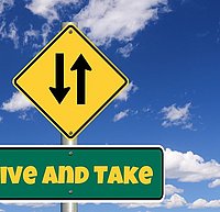 gelbes viereckiges Schild mit zwei entgegengesetzen Pfeilen vor einem blauen Himmel mit Wolken, darunter ein grünes Schild mit der Aufschrift in gelben Buchstaben "Give and Take"